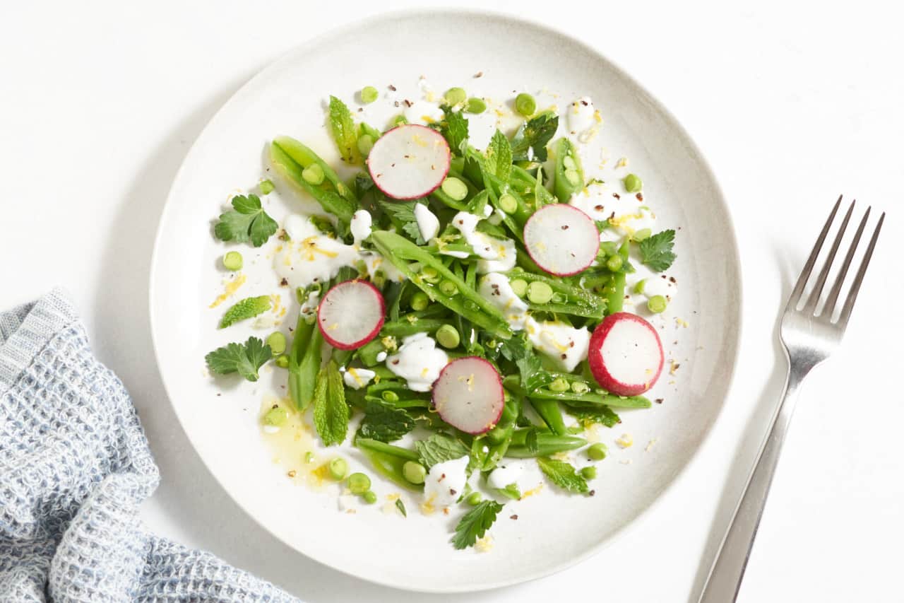 A plate of sugar snap pea salad topped with radishes and yogurt. A blue napkin is on the left, a fork is on the right.