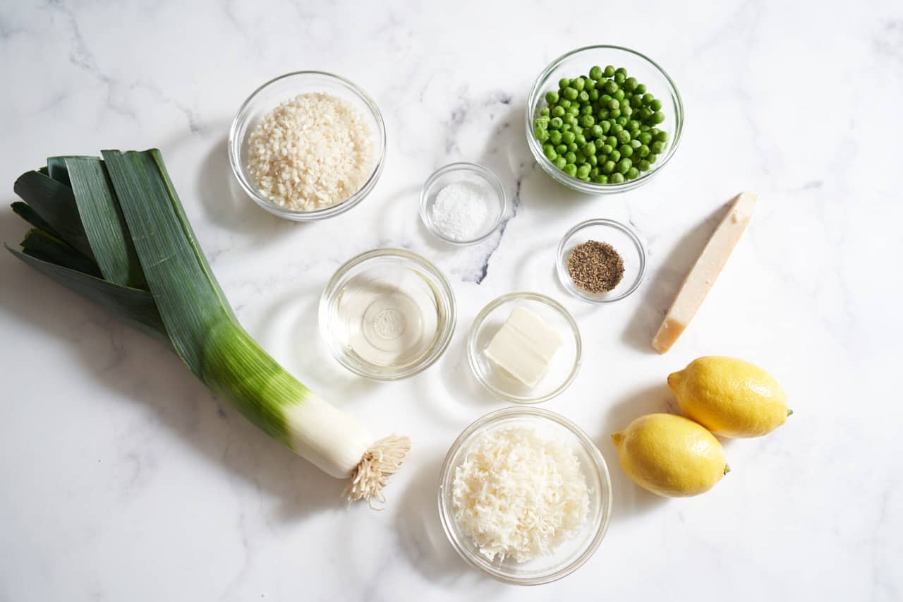 Two lemons, a leek, a parmesan rind, and small bowls of risotto rice, peas, grated parmesan, butter, white wine, salt, and pepper.