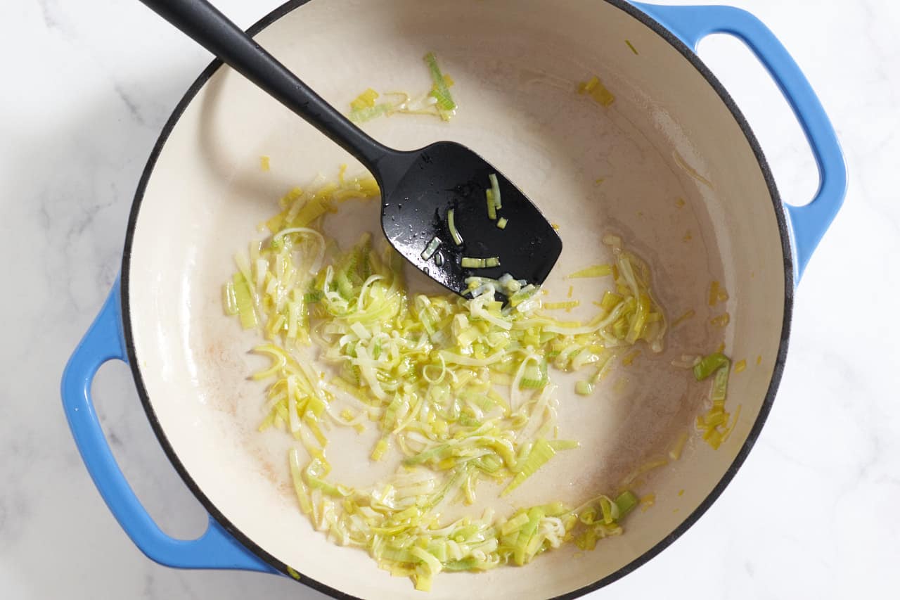 Softened leeks in a blue casserole pan with a black spatula.