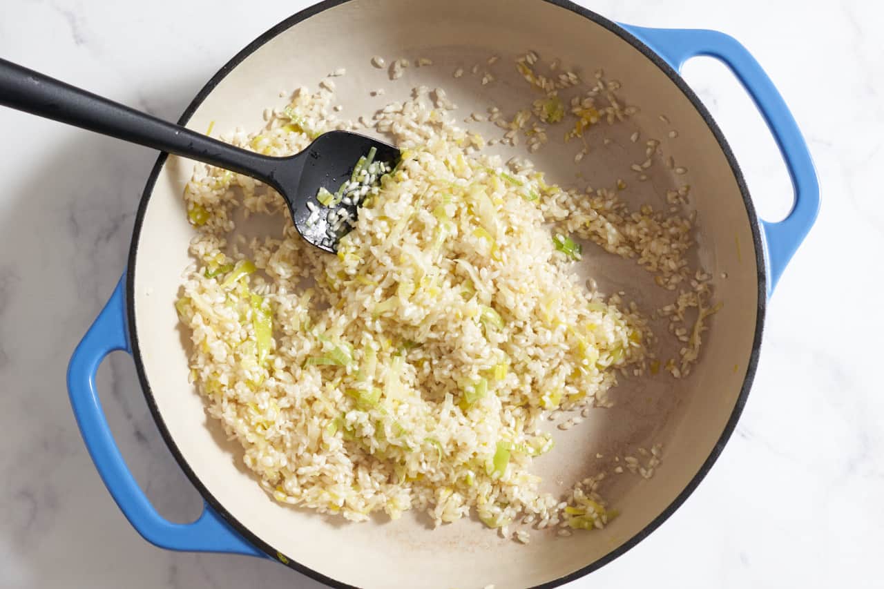 Risotto rice mixed with butter and leeks in a casserole pan with blue handles.