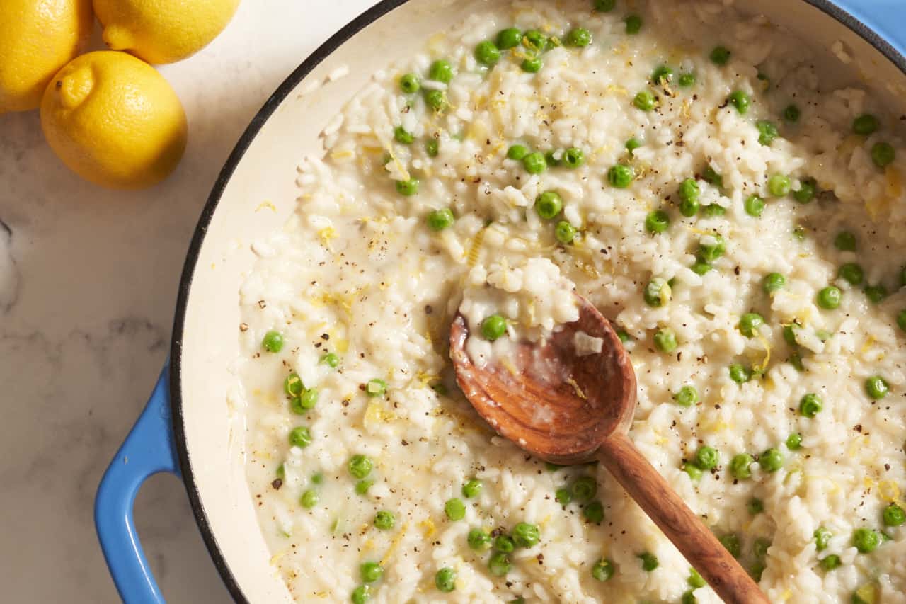 A pan of risotto with peas with a wooden spoon in it. Three lemons are in the upper left corner.