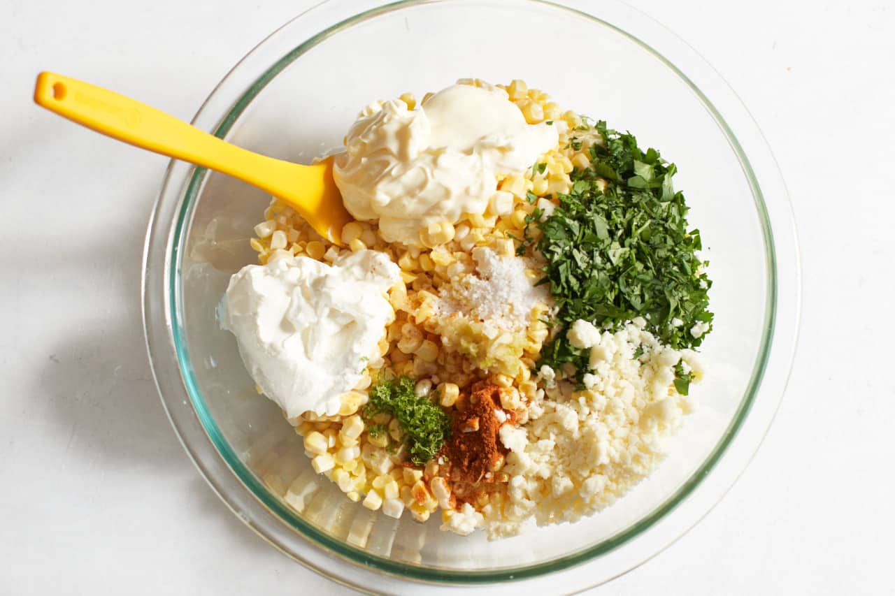 A yellow spatula in a bowl of fresh corn topped with sections of ingredients including mayonnaise, sour cream, cotija cheese, cilantro, and seasonings.