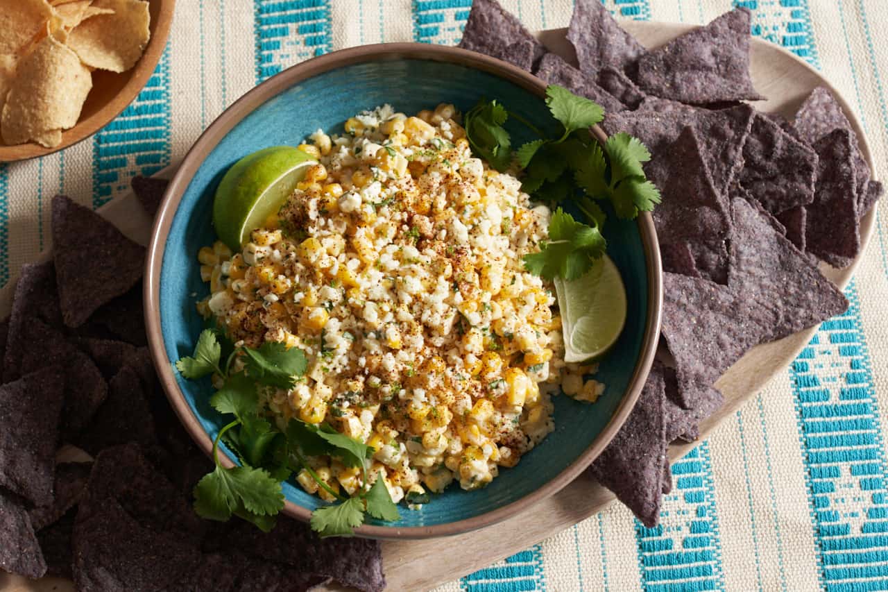 Elote dip in a blue bowl topped with cotija cheese and garnished with lime wedges and fresh cilantro. The bowl is on a plate of blue corn tortilla chips resting on a blue and white striped placemat, with a wooden bowl of yellow tortilla chips in the upper left.