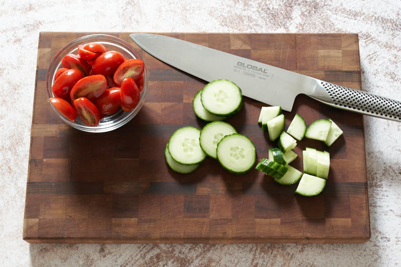 A knife on a cutting board with sliced cucumbers and a small bowl of sliced tomatoes.