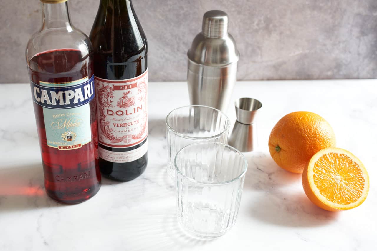 A bottle of Campari, a bottle of sweet vermouth, two empty rocks glasses, a cocktail shaker and jigger, an orange and half an orange.