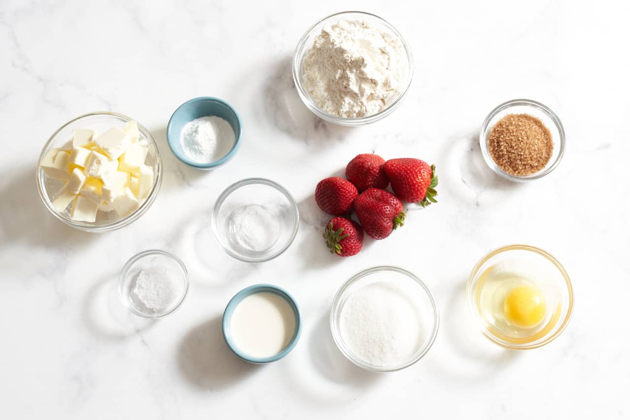 Fresh strawberries surrounded by small bowls of sugar, flour, butter, heavy cream, egg, baking powder, baking soda, and salt.