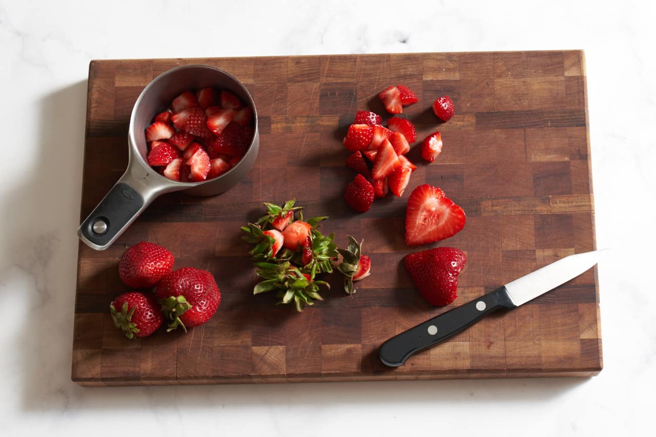 A knife on a cutting board with fresh strawberries that are being cut into small pieces.