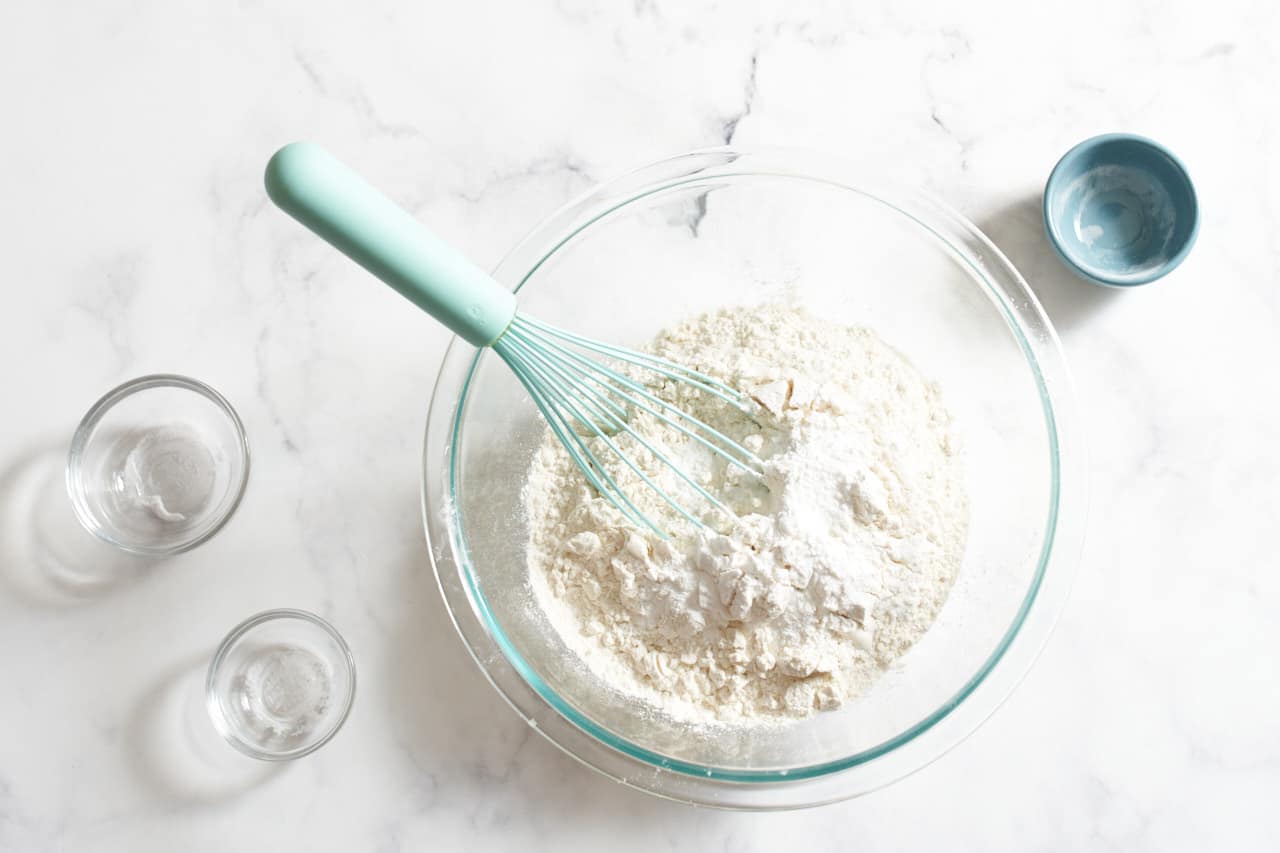 A whisk in a glass bowl of flour and other baking ingredients.