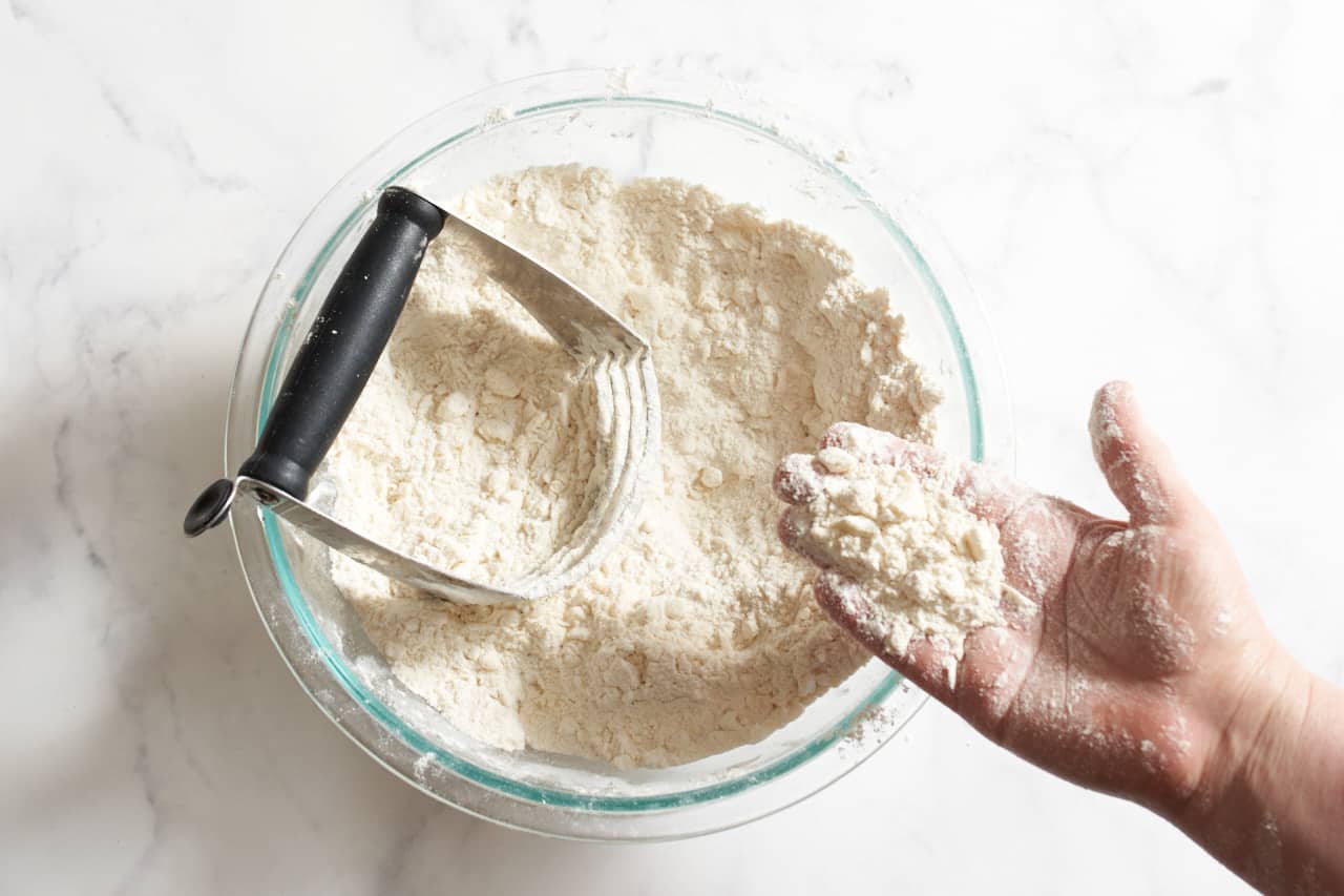 A woman's hand with small bits of butter that have been worked into flour. Her hand is over a bowl filled with flour, butter, and a pastry blender.