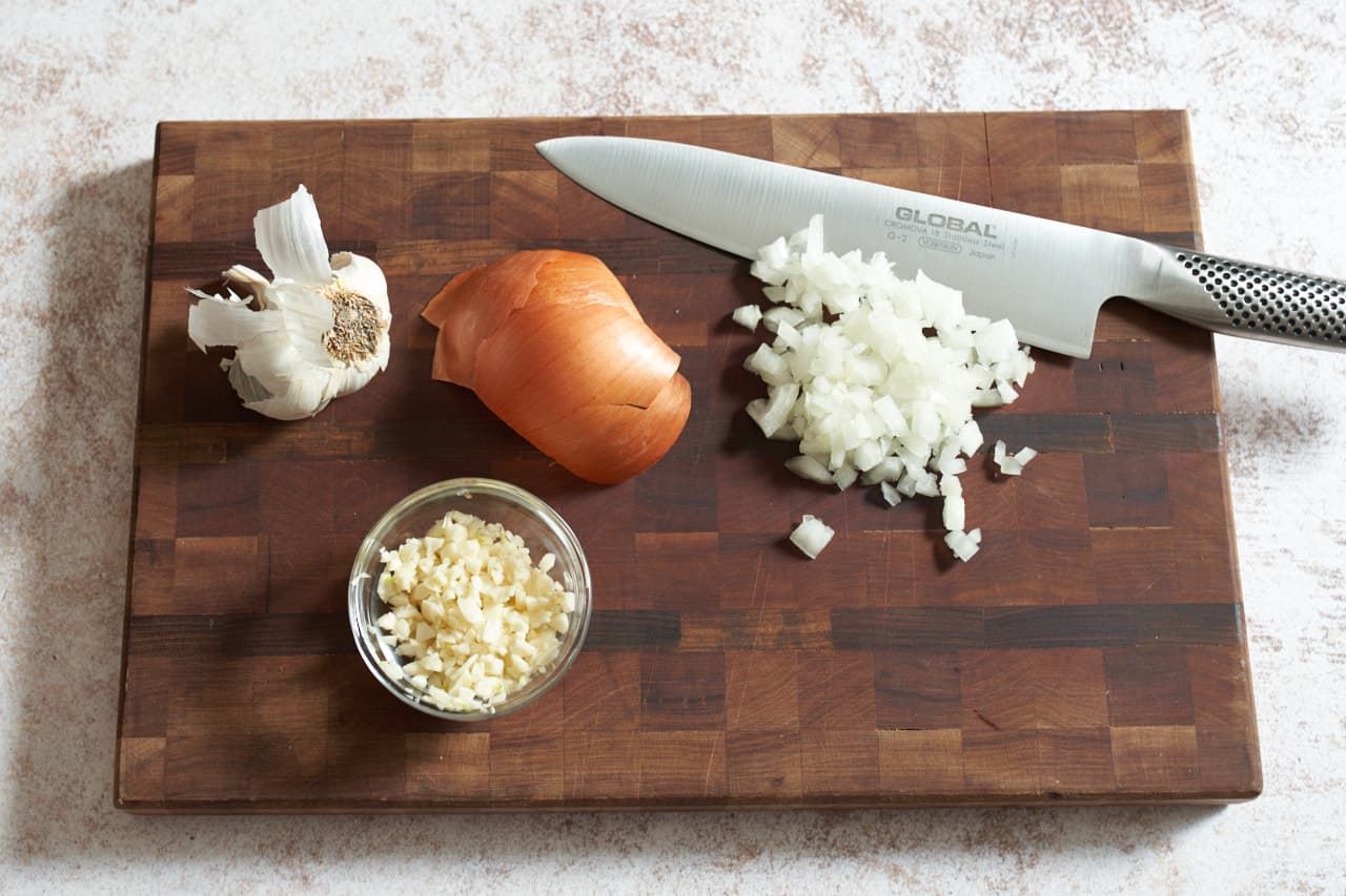 A knife on a cutting board with chopped onions and a bowl of chopped garlic.