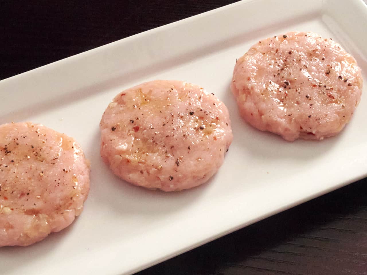 Three raw turkey burgers topped with salt, pepper, and olive oil on a white plate.