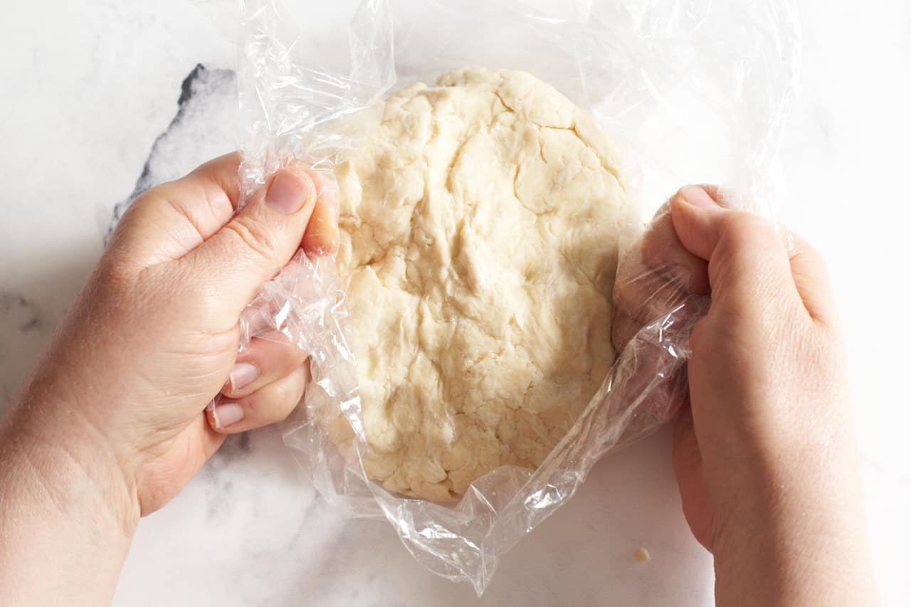 A woman's hands grasping plastic wrap that surrounds pie dough to demonstrate how to wrap it for chilling.