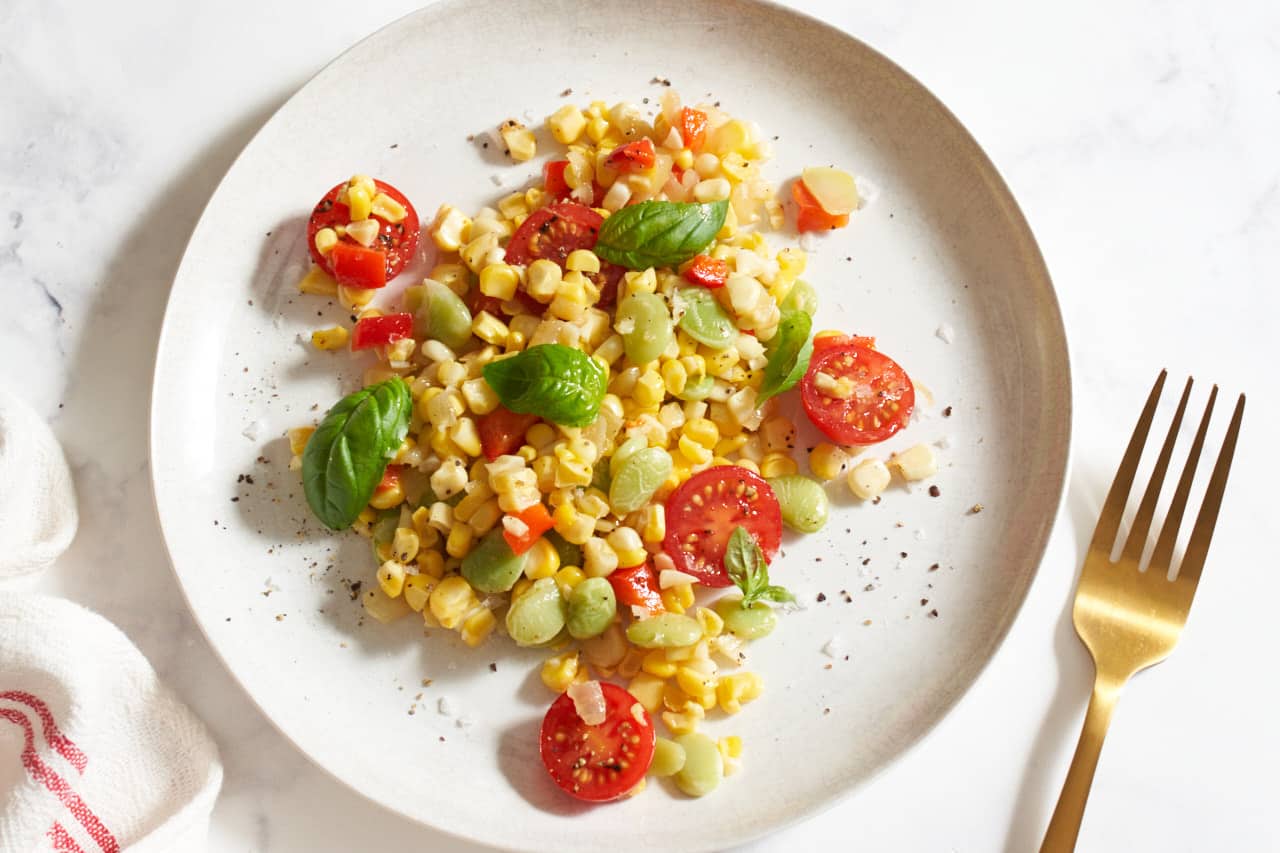 Corn succotash topped with fresh basil, salt, and pepper on a white plate. A gold fork is to the right, a red and white striped towel is on the left.