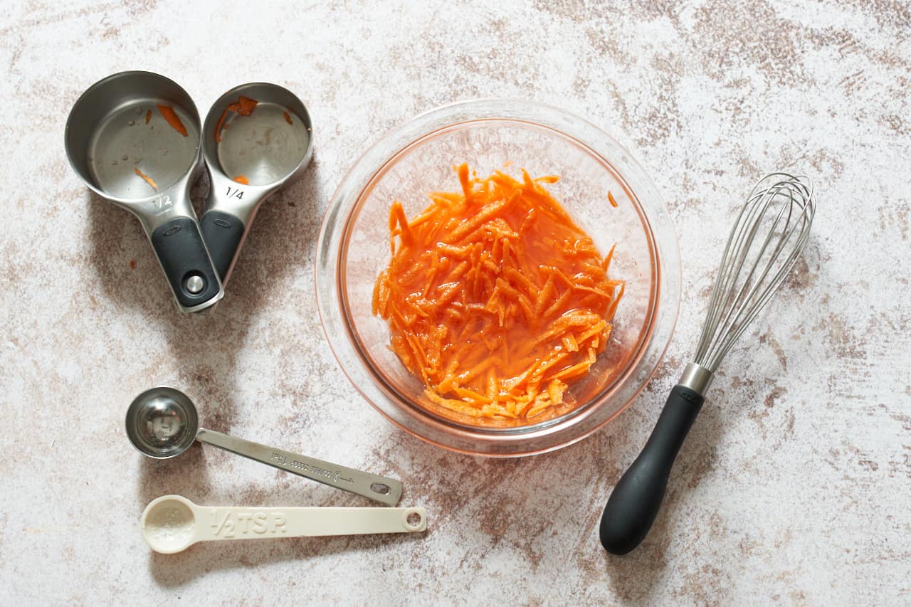 A bowl of grated carrots pickling in rice vinegar. A small whisk, two measuring cups, and two measuring spoons surround the bowl.