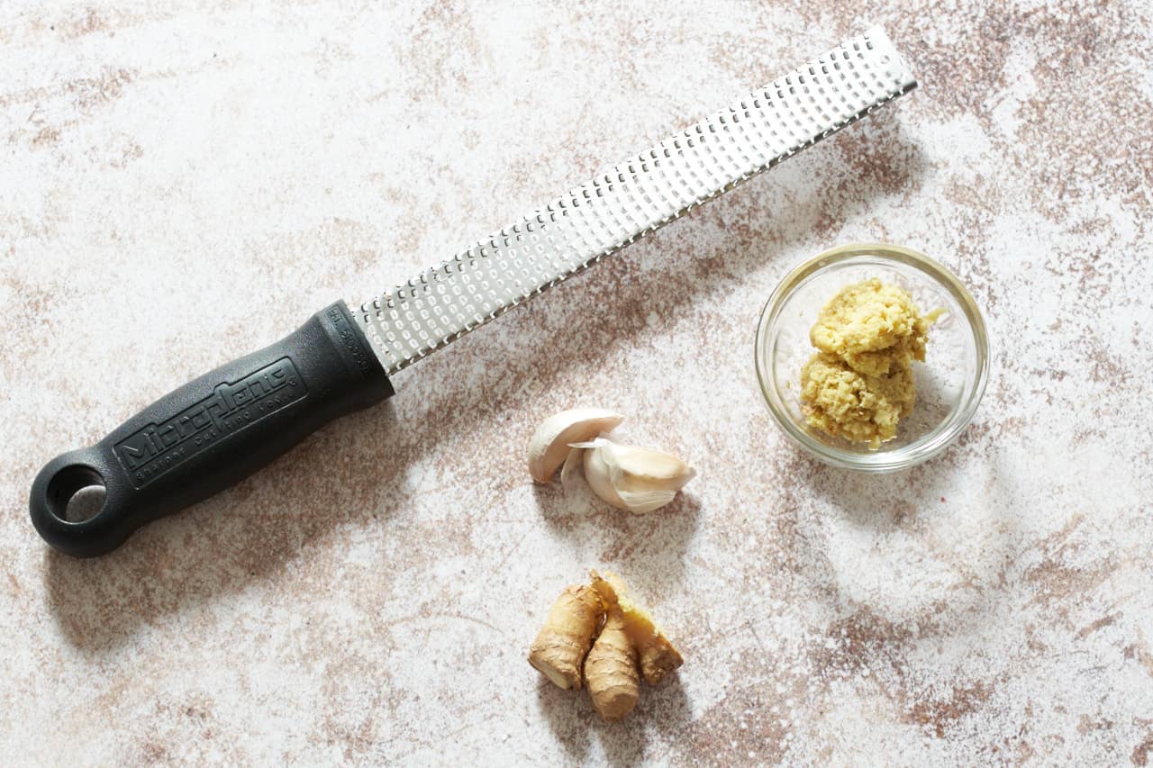 A microplane grater next to a small bowl of grated ginger, two garlic cloves, and a stem of ginger.