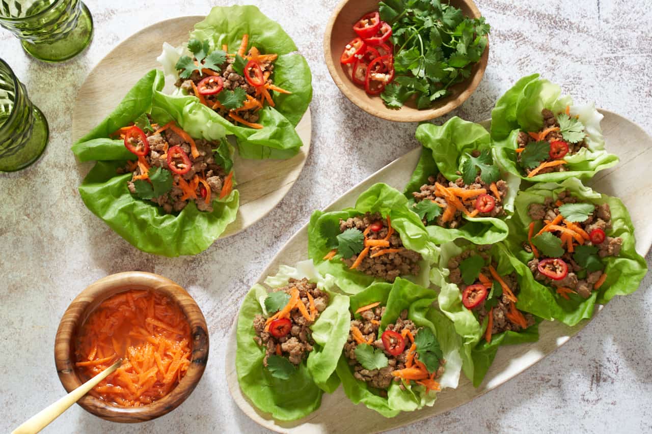 Pork lettuce wraps on an oval tray and on a small plate. Two green glasses are in the upper left, small wooden bowls in the bottom left and upper right have pickled carrots, fresh cilantro, and sliced red chilis.