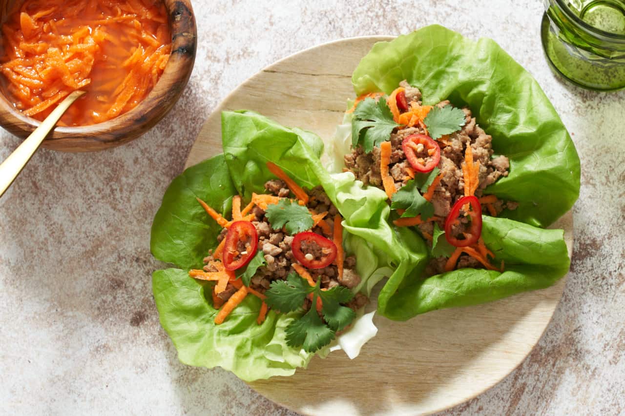A small bamboo plate with two pork lettuce wraps. A bowl of pickled carrots is on the left and a green glass is on the right.