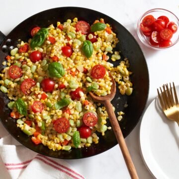 Succotash topped with fresh basil in a black skillet with a wooden spoon in it. A red and white towel is in the bottom left, a plate with two gold forks is on the right, and a small bowl of sliced cherry tomatoes is in the top right corner.