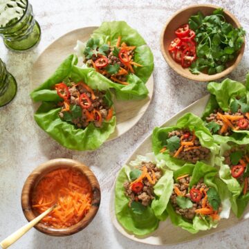 Pork lettuce wraps on an oval tray and on a small plate. Two green glasses are in the upper left, small wooden bowls in the bottom left and upper right have pickled carrots, fresh cilantro, and sliced red chilis.