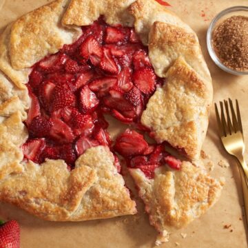 A strawberry galette with one slice cut into it. Two gold forks and a bowl of raw sugar are on the right. A strawberry is in the bottom left corner.