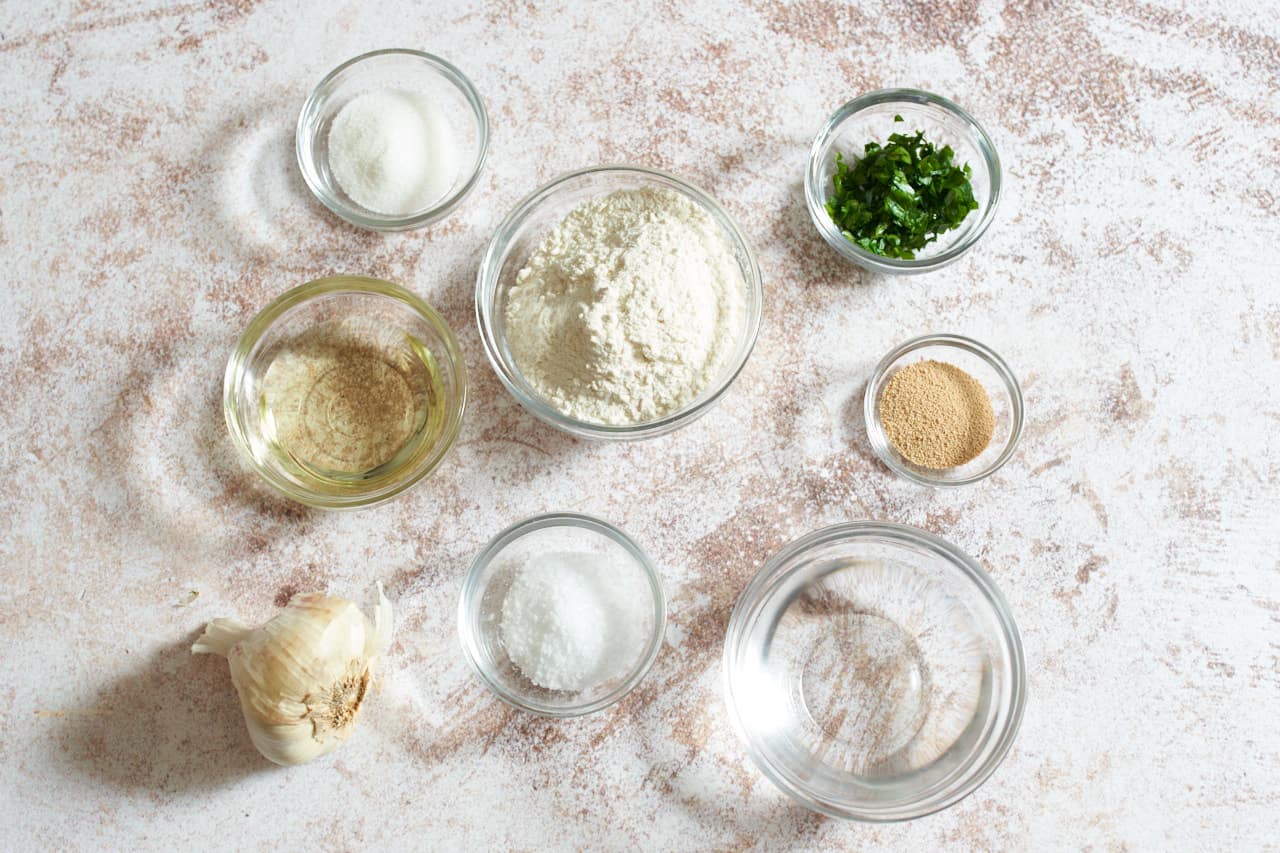A garlic bulb and small glass bowls of flour, sugar, salt, dry yeast, water, sunflower oil, and chopped parsley.