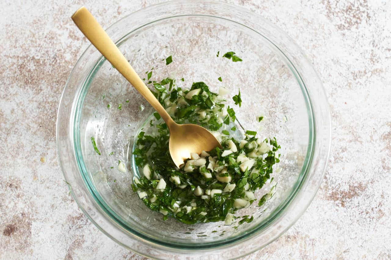 Chopped garlic, parsley, and sunflower oil in a glass bowl with a gold spoon in it.