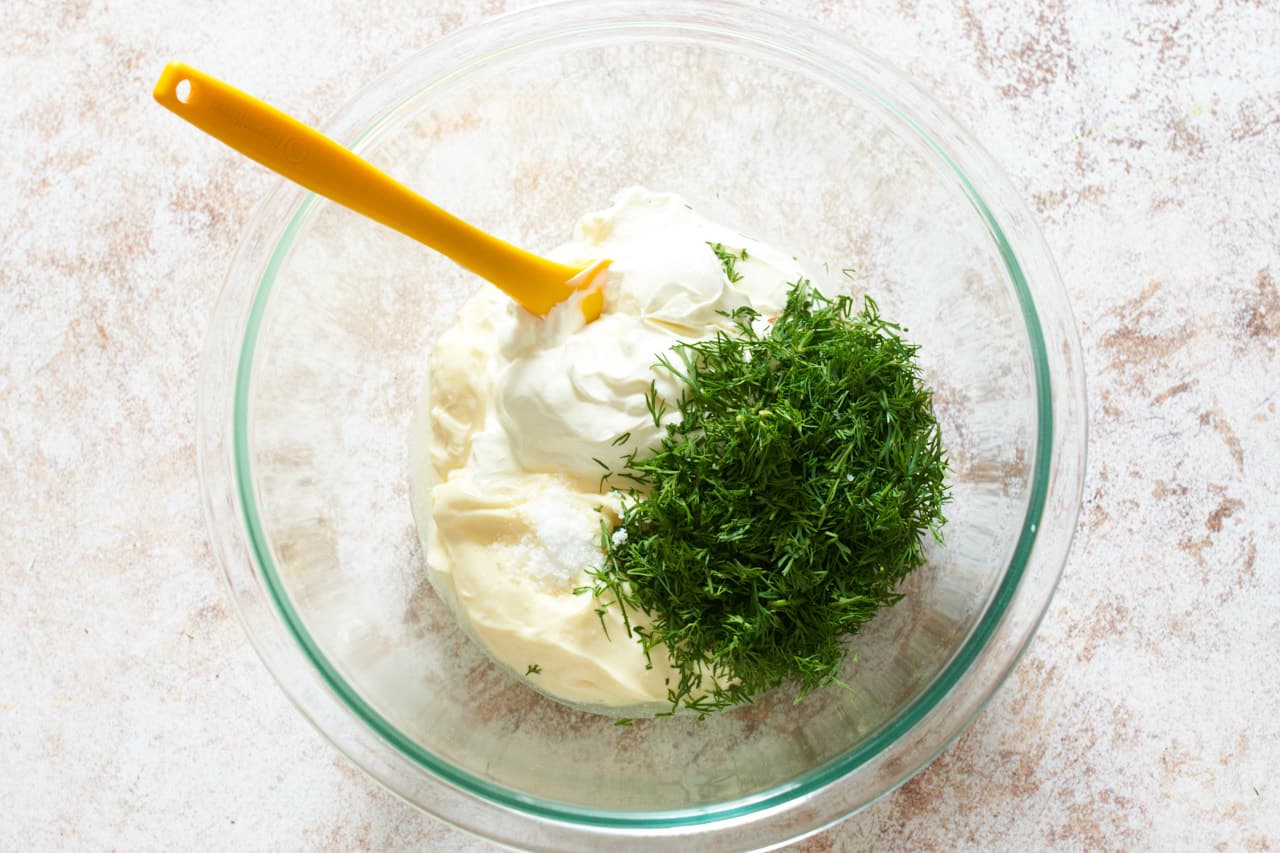 A mixing bowl of sour cream, mayonnaise, chopped dill, salt, and cider vinegar with a yellow spatula in it.