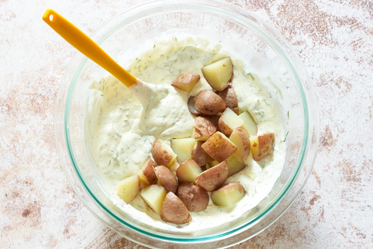 Red potatoes begin mixed into a creamy salad dressing in a glass bowl with a yellow spatula.