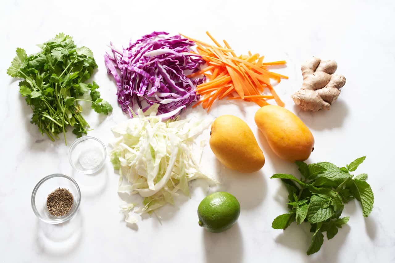 Sliced red and green cabbage, grated carrots, fresh cilantro, fresh mint, two mangoes, a lime, a stem of ginger and two small bowls of salt and pepper.