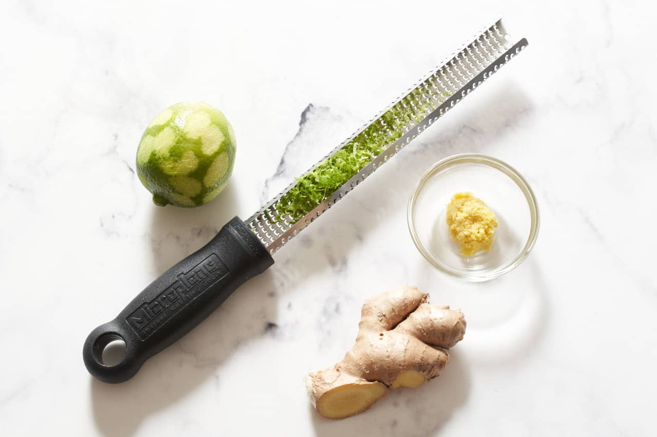 A lime that has been zested, a microplane grater with lime zest on it, a stem of ginger and a bowl of grated ginger.