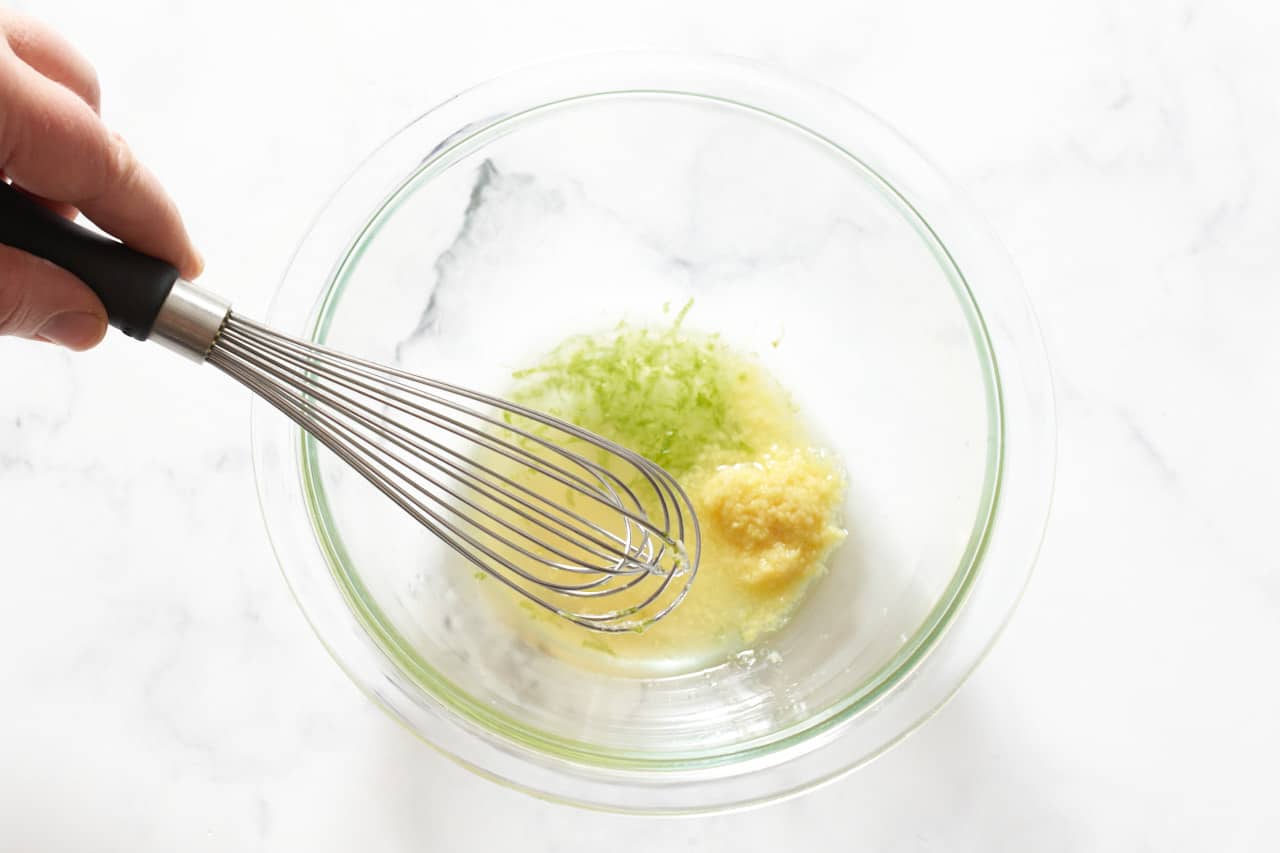 A woman's hand holding a whisk in a bowl of lime juice with zest and grated ginger.