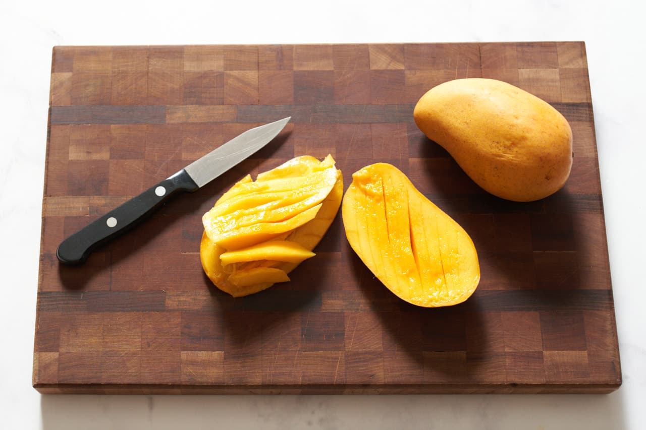 A whole mango and a mango that has been halved and cut into strips on a cutting board with a paring knife.