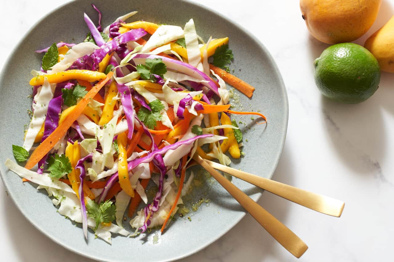 A plate of mango slaw with two gold utensils on it. Two mangoes and a lime are in the upper right corner.