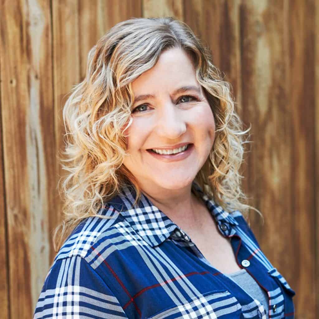 A woman with blond hair smiling at the camera. She is in front of a wooden fence and wearing a blue plaid shirt.