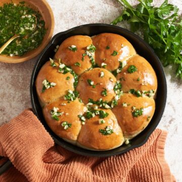 Pampushky (Ukranian garlic bread) in a cast iron skillet topped with a garlic-herb oil. A bowl of the oil is to the left, fresh parsley is on the right, the skillet handle is wrapped in an orange towel.