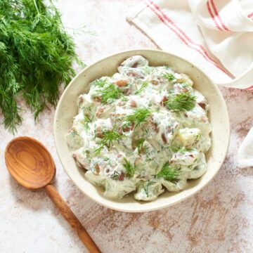 A bowl of dill potato salad topped with chopped fresh dill. A red and white striped towel, a wooden spoon, and fresh dill have been placed around the bowl.