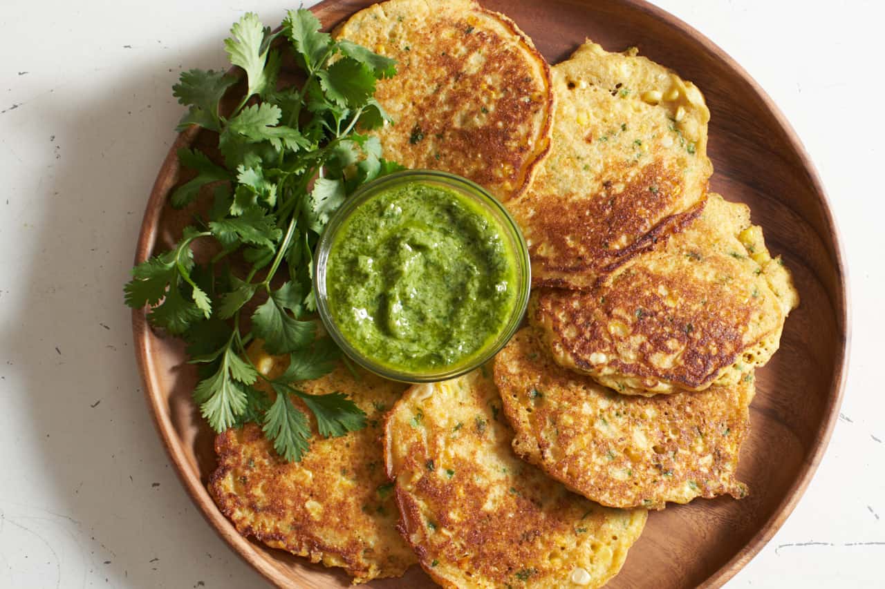 Corn cakes arranged in a circle on a wooden plate. A green sauce is in a small bowl in the center, and a fresh cilantro garnish is on the left side of the plate.