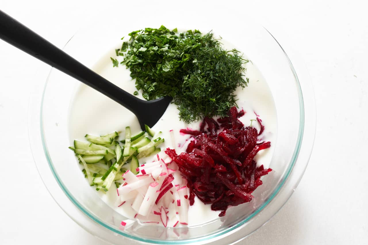 Chopped dill and parsley, and sliced beets, cucumber, and radishes being added to a bowl of kefir with a black spatula in it.