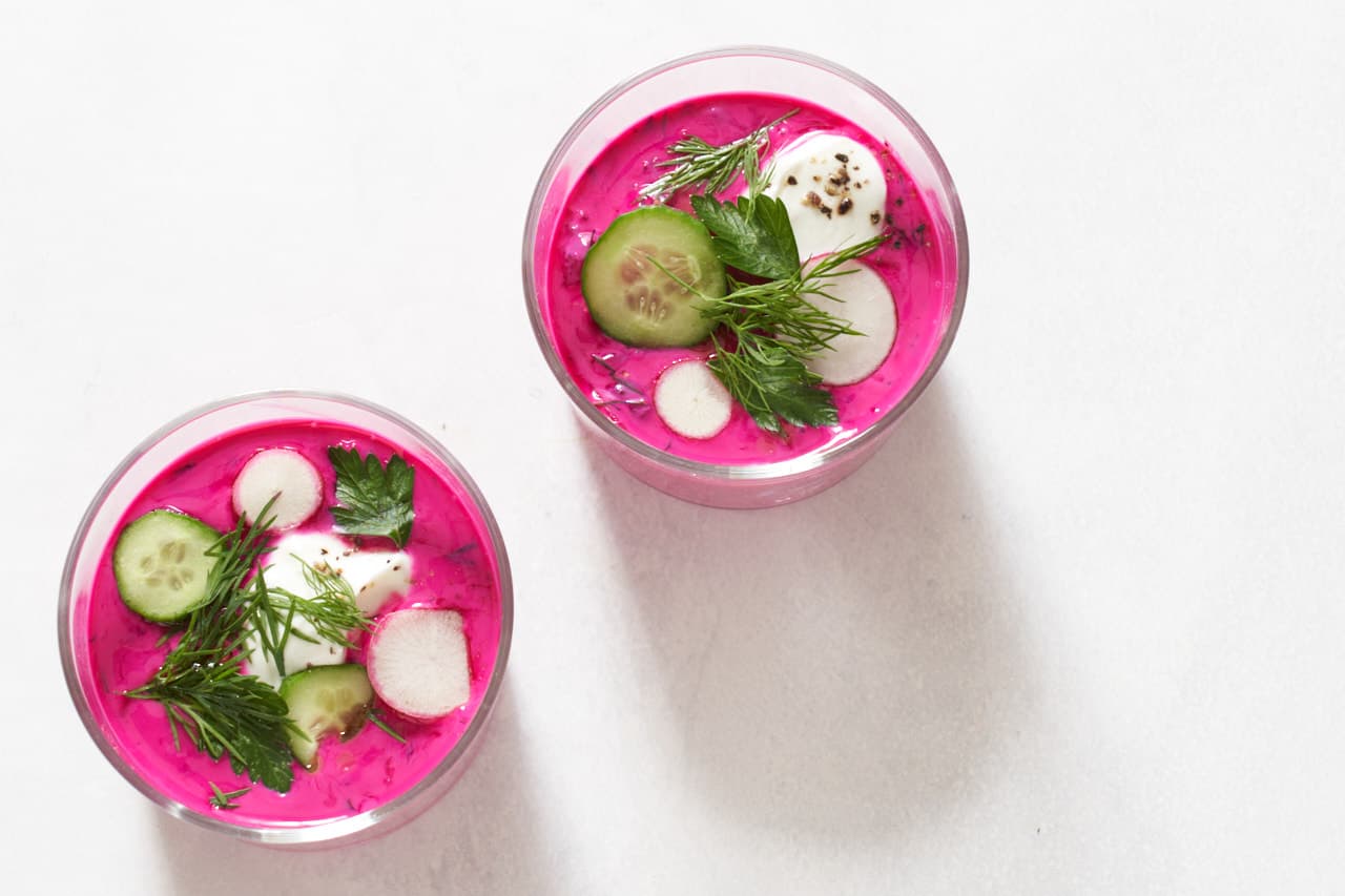 Two small glasses of cold borscht topped with sliced cucumbers and radishes, fresh herbs, and yogurt.