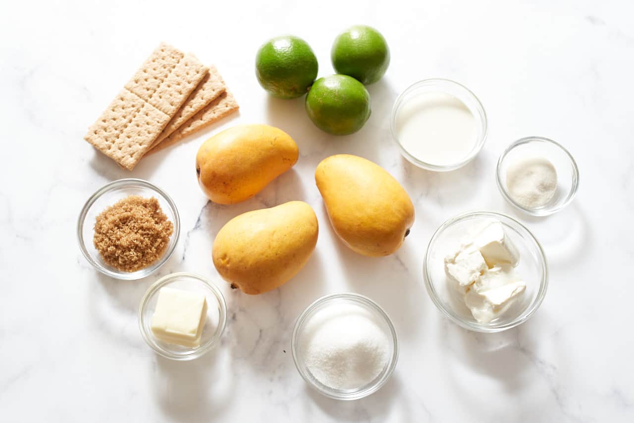 Ingredients for mango cheesecake: Three mangos, three limes, 3 graham crackers, and small glass bowls of brown sugar, butter, sugar, cream cheese, gelatin, and heavy cream.