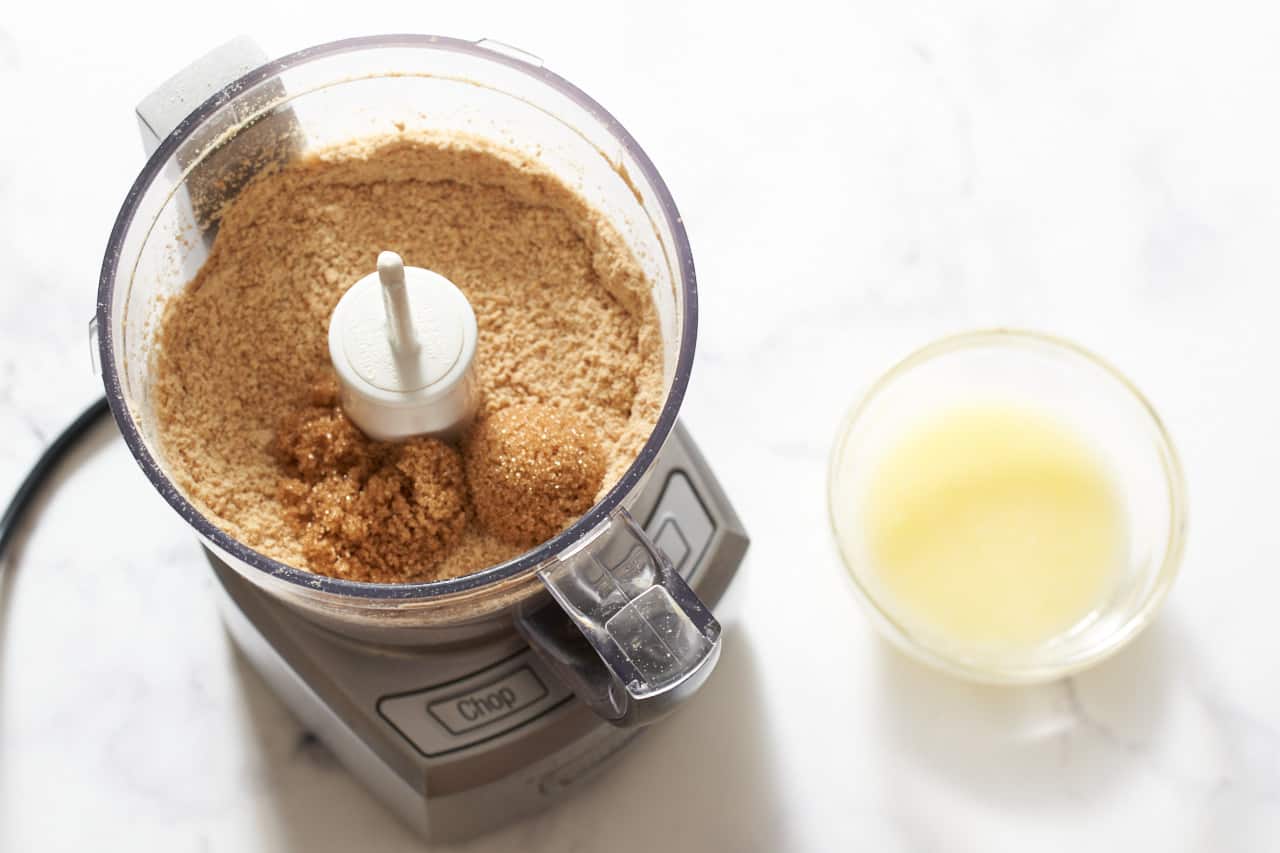 A small food processor with graham cracker crumbs and brown sugar next to a small bowl of melted butter.