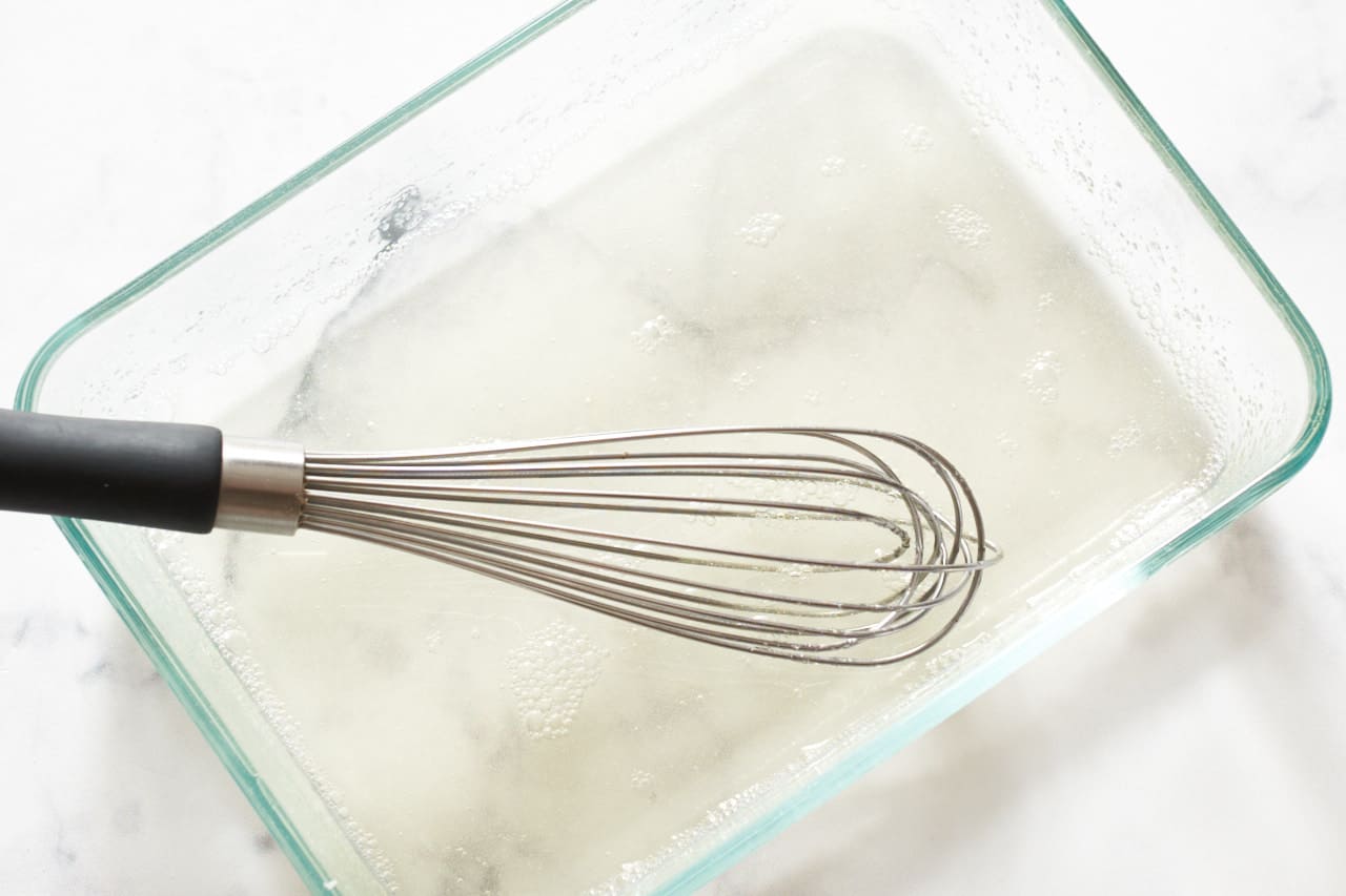 A small whisk in a rectangular glass container of clear liquid gelatin.