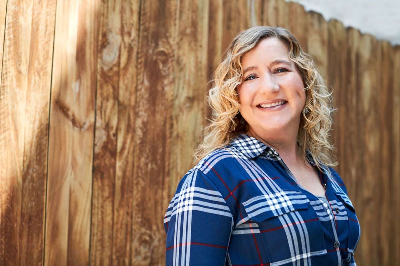 A woman with blond curly shoulder length hair wearing a blue plaid shirt, she is smiling and standing in front of a wooden fence.