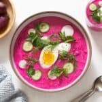 A bowl and a small glass of cold borscht with toppings including sliced cucumbers and radishes, fresh herbs, and a hard boiled egg. Fresh herbs, two spoons, a blue napkin, and a bowl of radishes surround the soups.