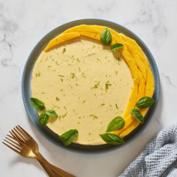 A no bake mango cheesecake topped with fresh mango slices arranged in a crescent moon shape, and fresh mint and basil leaves on a blue plate. There are three gold forks to the left and a blue napkin to the right of the plate.