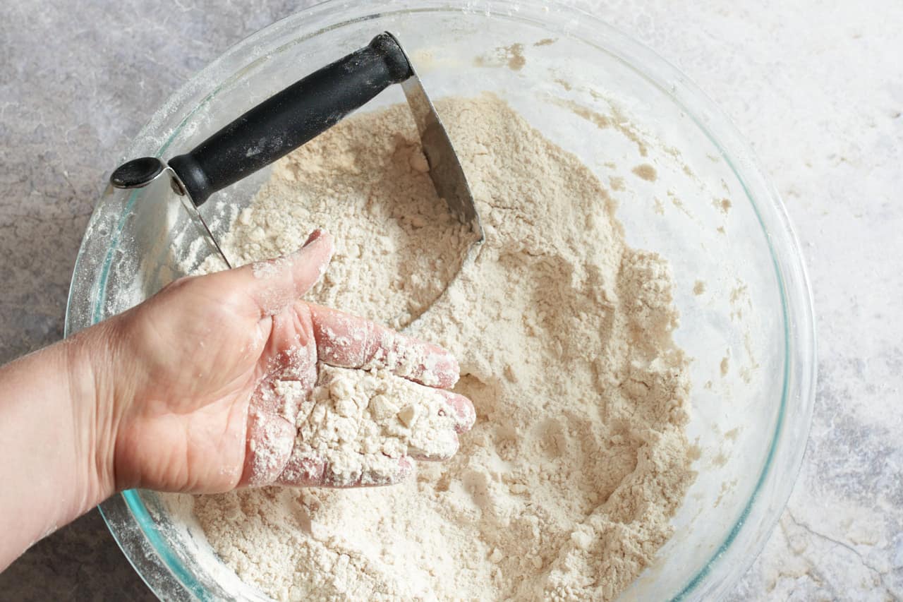 A woman's hand holding flour and butter that has been blended together over a bowl of flour with a pastry blender in it.