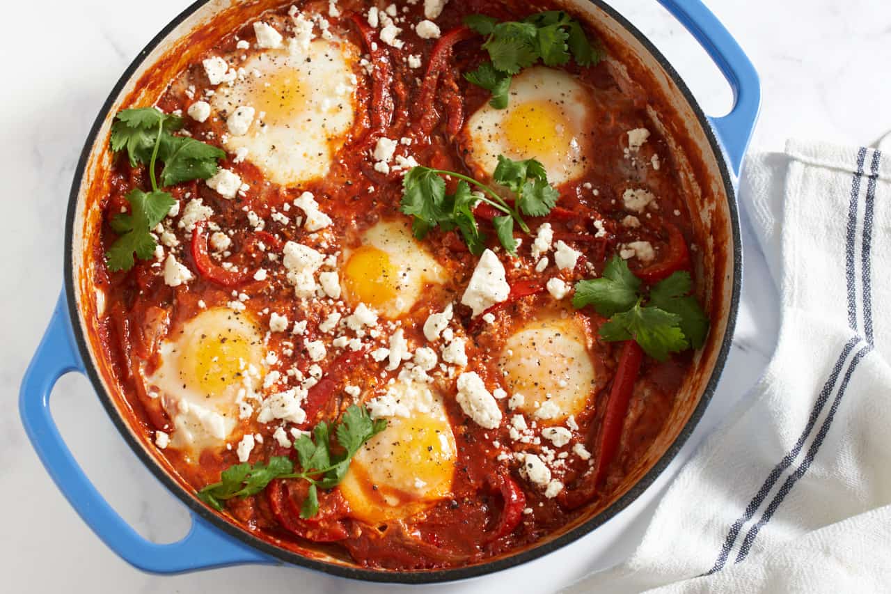 Shakshuka with feta and cilantro in a blue pan with a blue and white striped towel next to it.