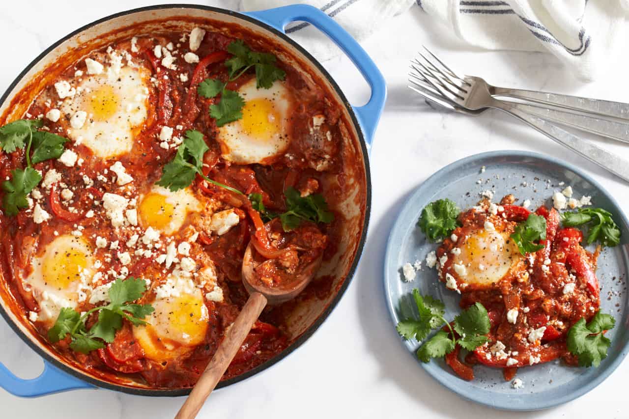 Shakshuka topped with feta and cilantro in a blue pan with a wooden spoon in it. A portion of shakshuka is on a blue plate to the right, forks and a blue and white striped towel are in the upper right corner.