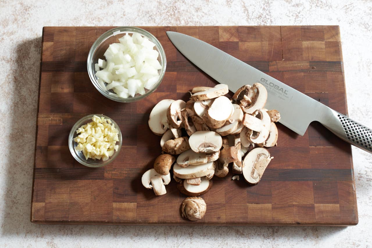 A knife on a cutting board with sliced mushrooms and two small bowls of diced onions and garlic.