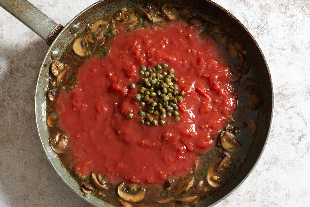 Tomatos and capers being added to a skillet of mushrooms and broth.